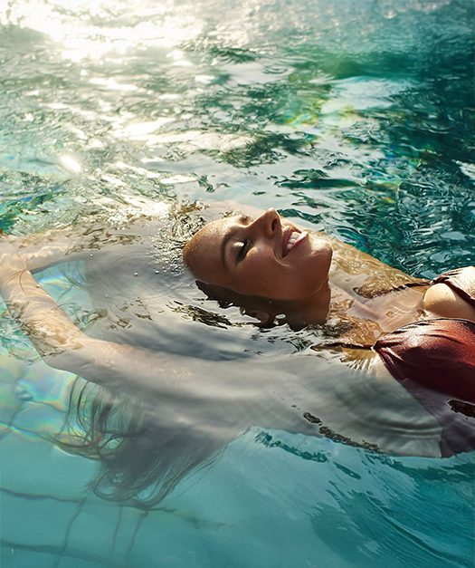 Woman in a swimsuit floating on her back in a sunlit turquoise pool.