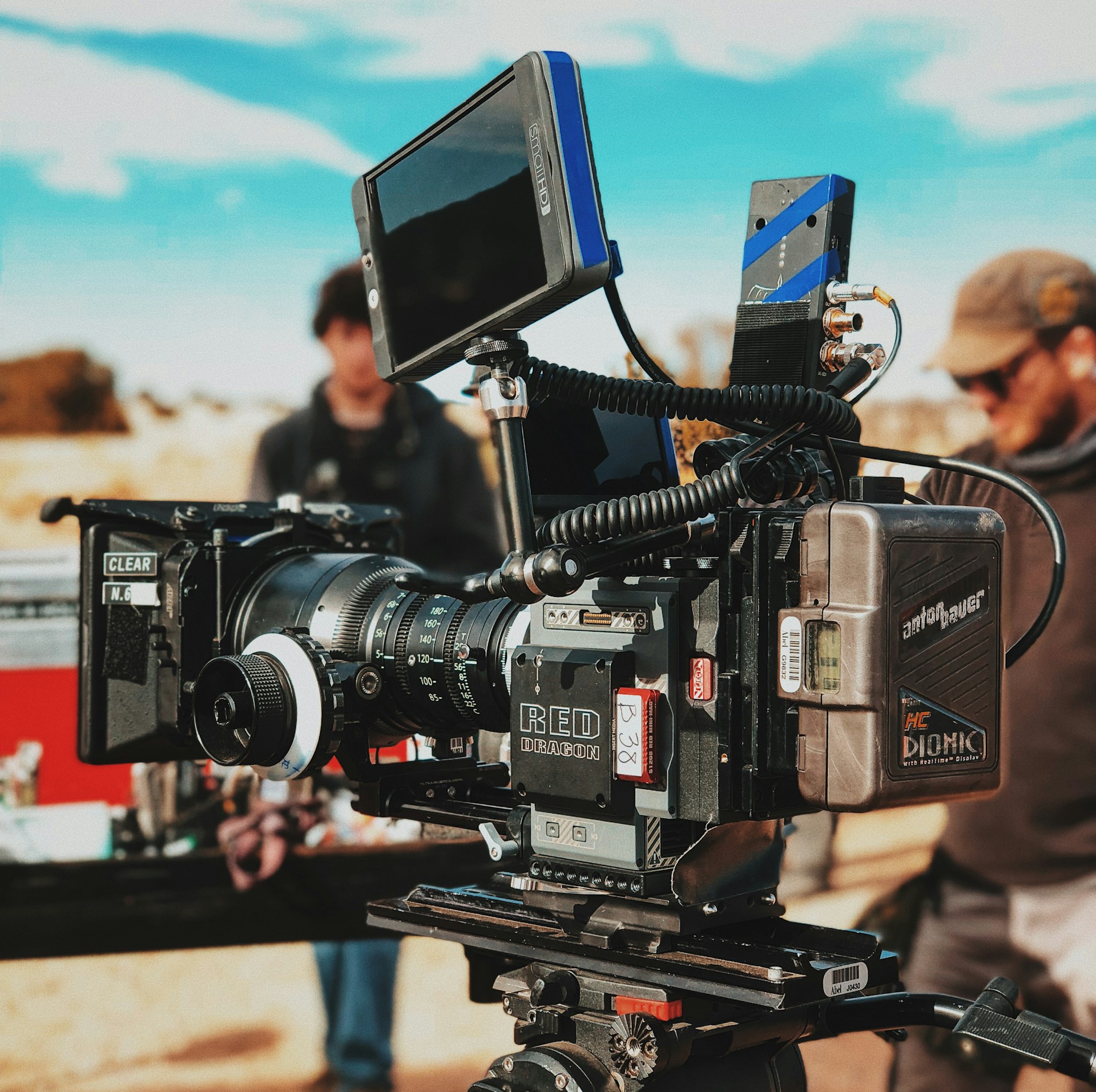 Professional RED Dragon cinema camera on a tripod outdoors with crew behind.