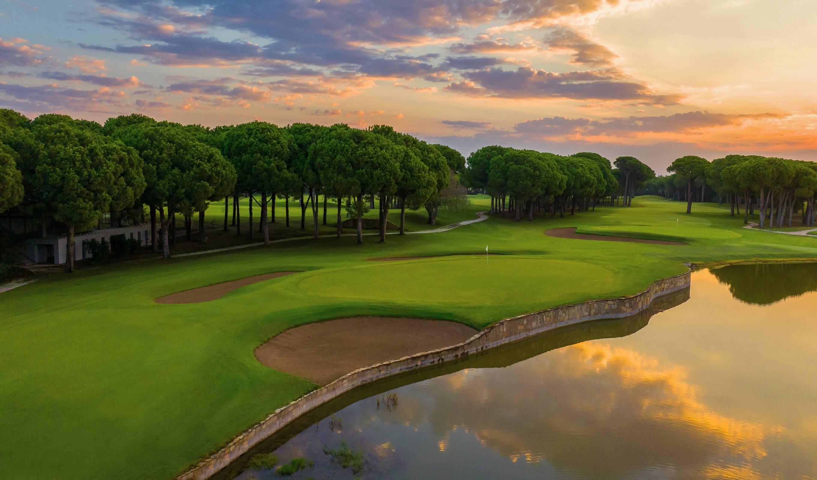 Gün batımında yeşil golf sahası, bunkerler ve göl manzarası.